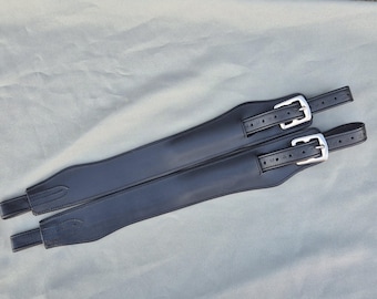 May include: A pair of black leather horse girth straps featuring contoured padding, reinforced stitching, and silver-toned metal buckles. The straps are designed with multiple adjustment holes and a smooth finish, showcasing a durable construction for equestrian use.