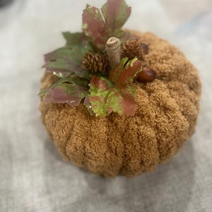 May include: A plush, brown pumpkin decoration with a textured surface. It is adorned with green and burgundy leaves, small pinecones, and acorns. The stem is wrapped in twine, creating a rustic autumn theme.
