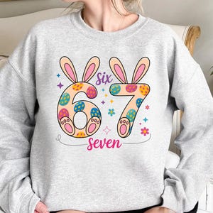 Pode incluir: Um moletom cinza claro com um design colorido de Páscoa. O design inclui os números "6" e "7" decorados com padrões de ovos de Páscoa e orelhas de coelho. A palavra "Six" está acima do "6" e "Seven" está abaixo do "7".