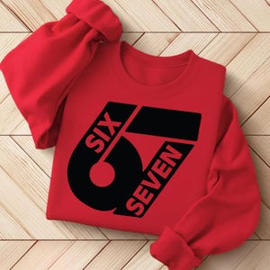 May include: A red sweatshirt with the text "SIX 67 SEVEN" in black. The graphic is a stylized design with the number 6 and the words SIX and SEVEN. The sweatshirt is made of a soft material and has long sleeves.