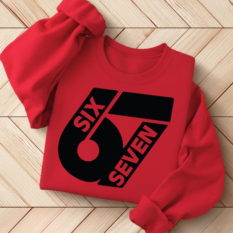 May include: A red sweatshirt with the text "SIX 67 SEVEN" in black. The graphic is a stylized design with the number 6 and the words SIX and SEVEN. The sweatshirt is made of a soft material and has long sleeves.