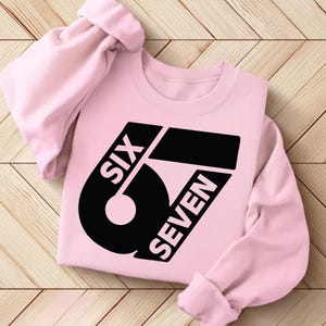 May include: A light pink sweatshirt with the words "SIX SEVEN" in a bold black font. The design features a large number "67" with the words arranged around it. The sleeves are rolled up, and the sweatshirt is lying on a wooden surface.