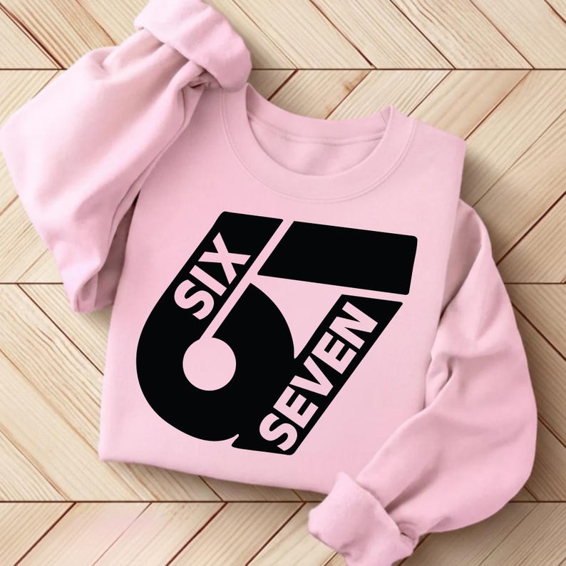 May include: A light pink sweatshirt with the words "SIX SEVEN" in a bold black font. The design features a large number "67" with the words arranged around it. The sleeves are rolled up, and the sweatshirt is lying on a wooden surface.