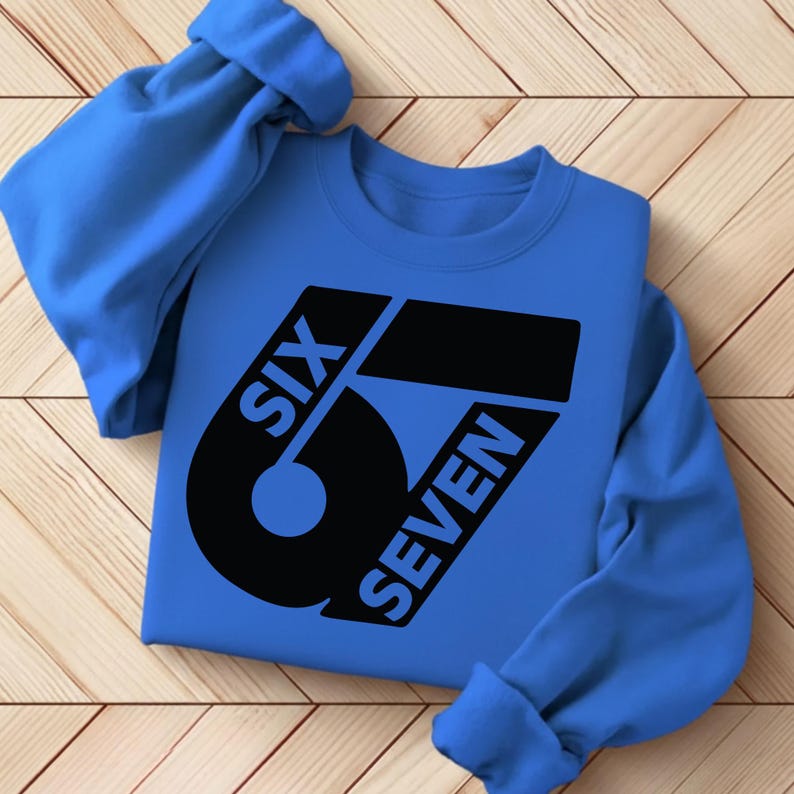 May include: A blue sweatshirt with the words "SIX SEVEN" in a black, stylized font. The design is centered on the front of the sweatshirt. The sleeves are rolled up, and the sweatshirt is lying on a wooden surface.
