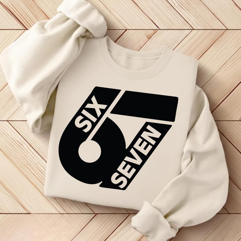 May include: Cream-colored sweatshirt with the words "SIX SEVEN" in a bold, black, stylized font. The design features a large number 6 and a horizontal bar. The sleeves are folded, and the sweatshirt is on a wooden surface.