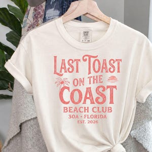 May include: An off-white short-sleeve t-shirt featuring red text that reads Last Toast on the Coast Beach Club 30A Florida Est. 2026, accompanied by a small graphic of a palm tree and a sun. The shirt is displayed on a wooden hanger.