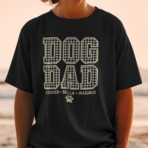 May include: Black t-shirt with the text "DOG DAD" in a houndstooth pattern. Below the text are the names "COOPER • BELLA • MAXIMUS" and a paw print. The shirt is a casual style.