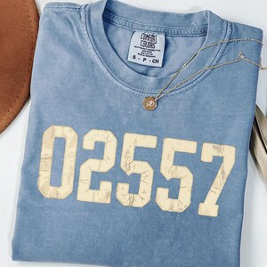May include: A blue short-sleeved t-shirt with a crew neckline featuring the number 02557 printed in a distressed cream font across the chest. The shirt has a Comfort Colors brand label inside the collar and is size small.