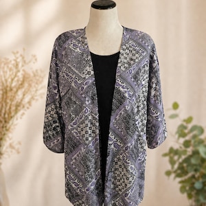 May include: An open-front kimono-style cardigan featuring a patchwork pattern in shades of purple, black, and white. The garment has three-quarter length sleeves and is layered over a solid black sleeveless top on a mannequin display.