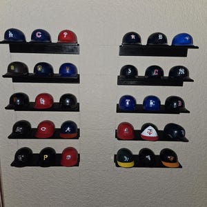 Könnte beinhalten: Sammlung von Miniatur-Baseballkappen, die auf schwarzen Regalen ausgestellt sind. Die Kappen zeigen verschiedene Teamlogos und -farben, darunter Blau, Rot und Schwarz. Die Anordnung ist symmetrisch und zeigt eine Vielzahl von Teamzugehörigkeiten.