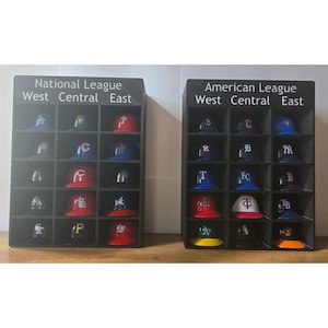 以下が含まれることがあります： 野球帽が並んだ2つの黒いディスプレイケース。1つは「ナショナルリーグ」、もう1つは「アメリカンリーグ」と表示されています。各ケースは「ウェスト、セントラル、イースト」とラベル付けされたセクションに分かれています。帽子はさまざまな色で、チームのロゴが特徴です。