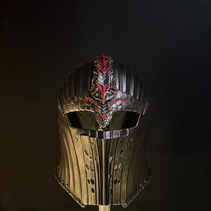 May include: A detailed, dark metal helmet with a red and silver crest. The helmet has a visor and a neck guard with vertical slots. The design suggests a medieval or fantasy theme, possibly for cosplay or display.