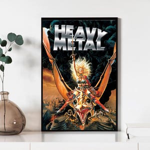 May include: A framed poster featuring the words "HEAVY METAL" in silver. The image shows a woman with blonde hair riding a winged beast, holding a sword aloft. The background includes a dark sky and a distant cityscape.