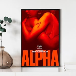 Puede incluir: Póster de película enmarcado con borde negro. El póster presenta una combinación de colores rojo y naranja con el título "ALPHA" en letras naranjas grandes. El póster también incluye el texto "A FILM BY JULIA DUCOURNAU" y el logotipo NEON.