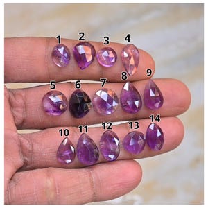 Puede incluir: Una colección de catorce piedras preciosas facetadas en varias formas y tonos de púrpura y rosa. Las piedras preciosas están numeradas y se muestran sobre un fondo neutro. Las piedras son de forma ovalada, pera y redonda.