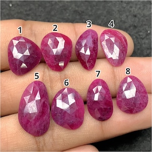 Puede incluir: Ocho piedras preciosas facetadas en forma de pera, de color rojo rosado intenso, dispuestas en dos filas. Cada piedra está numerada del uno al ocho en texto blanco. Las piedras presentan un corte tipo rosa con múltiples facetas.