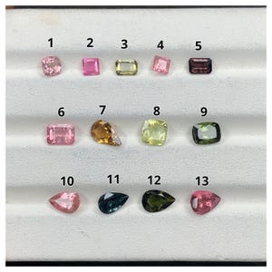 May include: Thirteen faceted loose gemstones arranged in three rows on a white display tray. The stones include various shapes like cushion, emerald, and pear cuts in colors ranging from light pink, yellow, and green to dark blue and purple.