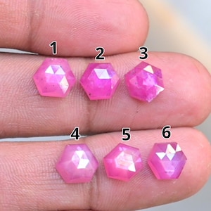 May include: Six faceted, hexagonal pink gemstones are displayed. The gems are a vibrant shade of pink, with multiple facets that catch the light. The gemstones are held in a hand, showcasing their size and shape. These could be used for jewelry making.