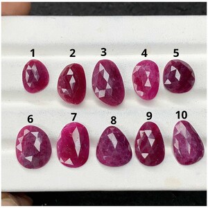 May include: Ten faceted, deep pink to red gemstones arranged in two rows on a white display tray. Each stone is numbered from 1 to 10 in black text above it. The gems feature various irregular, rounded shapes with geometric, multi-faceted surfaces.