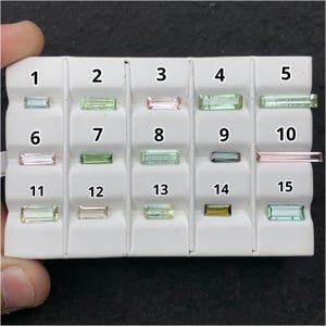 May include: A white display tray containing fifteen rectangular baguette-cut gemstones arranged in three rows of five. Each stone is numbered from 1 to 15 and features various colors including light green, pink, olive, and blue-green hues.