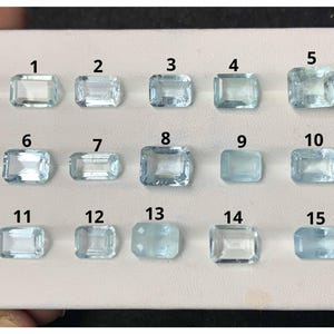 Pode incluir: Uma coleção de quinze pedras preciosas de água-marinha, cada uma com formato retangular ou quadrado. As pedras variam em tamanho e clareza, com um tom azul pálido. As pedras preciosas estão dispostas em uma superfície branca, numeradas de 1 a 15.