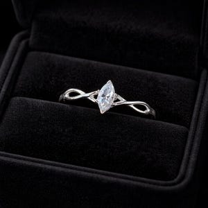 May include: A silver ring featuring a marquise-cut gemstone, set within a delicate, swirling design. The ring is presented in a black velvet ring box, accentuating its elegant appearance. The gemstone is clear and sparkling.