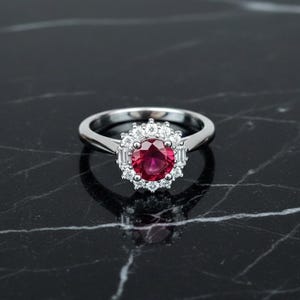 Puede incluir: Un anillo plateado con una gema roja oscura redonda y grande, rodeada de gemas claras más pequeñas. El anillo tiene una banda pulida y un diseño de halo. El anillo se muestra sobre una superficie de mármol negro.