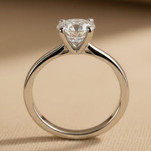 May include: A silver engagement ring featuring a large, clear, round-cut diamond. The ring has a simple, polished band and a four-prong setting. The ring is set against a neutral background. The ring's band is made of silver.