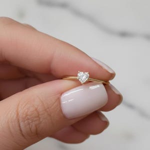 Puede incluir: Un delicado anillo de oro con un diamante en forma de corazón. El anillo tiene una banda delgada y una pequeña gema brillante. El anillo se sostiene sobre un fondo suave y neutro, resaltando el elegante diseño de la joya.