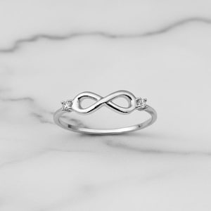 Op de afbeelding: Een zilverkleurige ring met een oneindigheidssymbool. De ring heeft een gepolijste afwerking en kleine, heldere edelstenen aan weerszijden van het oneindigheidssymbool. De ring staat tegen een witte marmeren achtergrond.