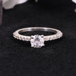 May include: A silver ring featuring a large, round, clear gemstone as the focal point. The band is adorned with smaller, clear gemstones. The ring is displayed against a black backdrop, accentuating its brilliance. A timeless piece of jewellery.