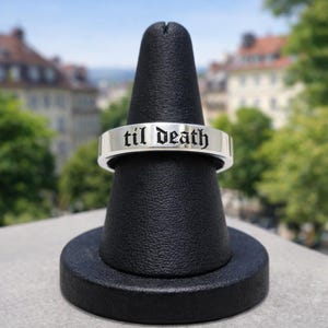 May include: A silver ring with the phrase "til death" engraved in a gothic font. The ring is displayed on a black ring holder against a blurred background of buildings and trees.