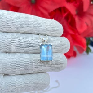 May include: A silver pendant necklace featuring a rectangular, light blue gemstone. The gemstone is faceted, catching the light. The pendant hangs from a delicate silver chain. The background includes a white surface and red flowers.