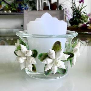 May include: A clear glass candle holder with a white candle inside. The holder is decorated with white seashells and green accents. The candle holder is a decorative item.