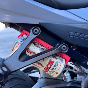 Puede incluir: Primer plano de la suspensión trasera de una motocicleta con un soporte rojo que sujeta dos latas de Monster Energy Juice. Las latas tienen un fondo beige con gráficos y texto en rojo y negro.