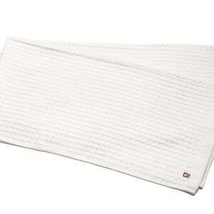 May include: A folded white waffle-weave towel. The towel has a textured surface and a small red and blue logo in the corner. The towel is made of a soft material and is ideal for use in a bathroom or spa.