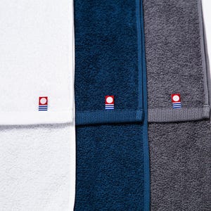 Puede incluir: Primer plano de tres toallas dobladas en blanco, azul marino y gris. Cada toalla tiene un pequeño emblema bordado con un cuadrado rojo, un círculo blanco y rayas azules. Las toallas están hechas de un material suave y absorbente.
