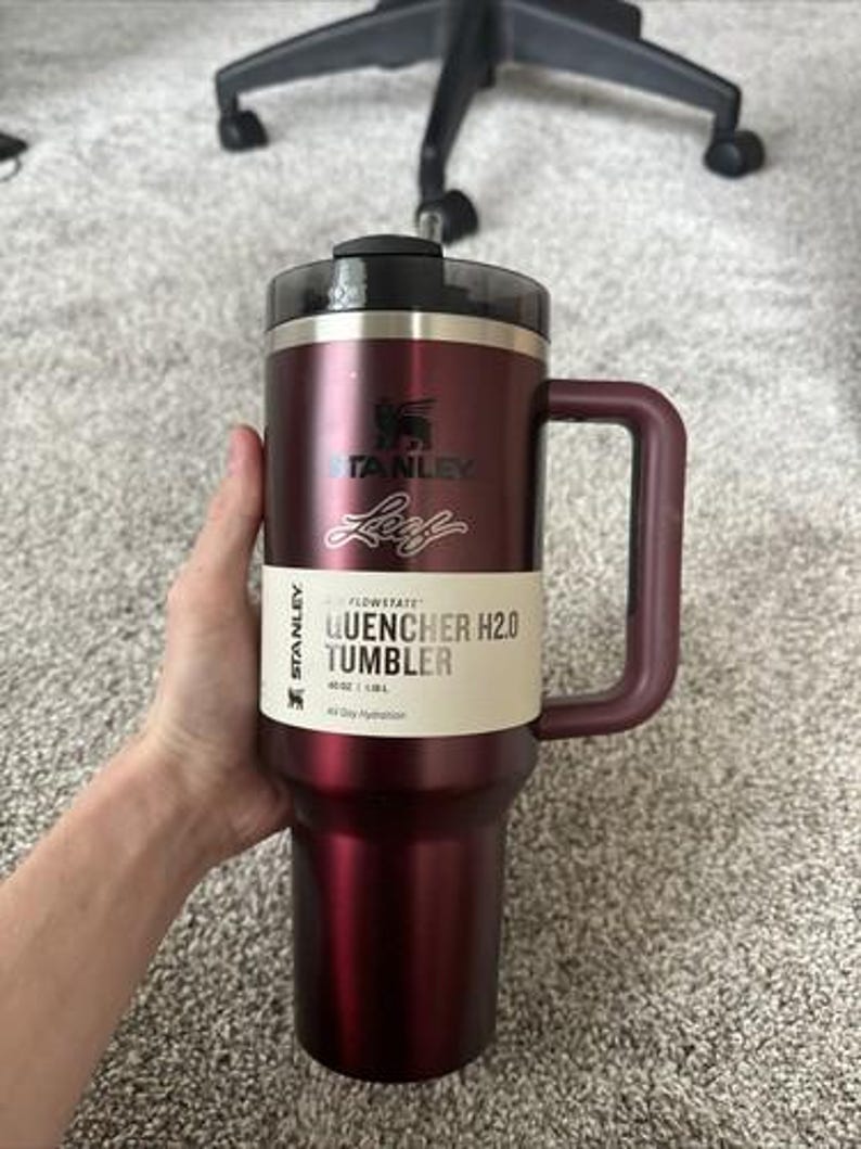 May include: A burgundy Stanley Quencher H2.0 Tumbler with a handle. The tumbler has a black lid and a white label with the text "QUENCHER H2.0 TUMBLER". The tumbler is designed for all-day hydration and holds 40 oz / 1.18L.