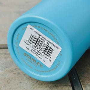 May include: A light blue Stanley 40oz tumbler. The base of the tumbler features a white sticker with product details, including the text "STAN ADV 40OZ QNCHRH2.0 TM TUMB POOL (BOX)" and a barcode. The tumbler is BPA free and dishwasher safe.