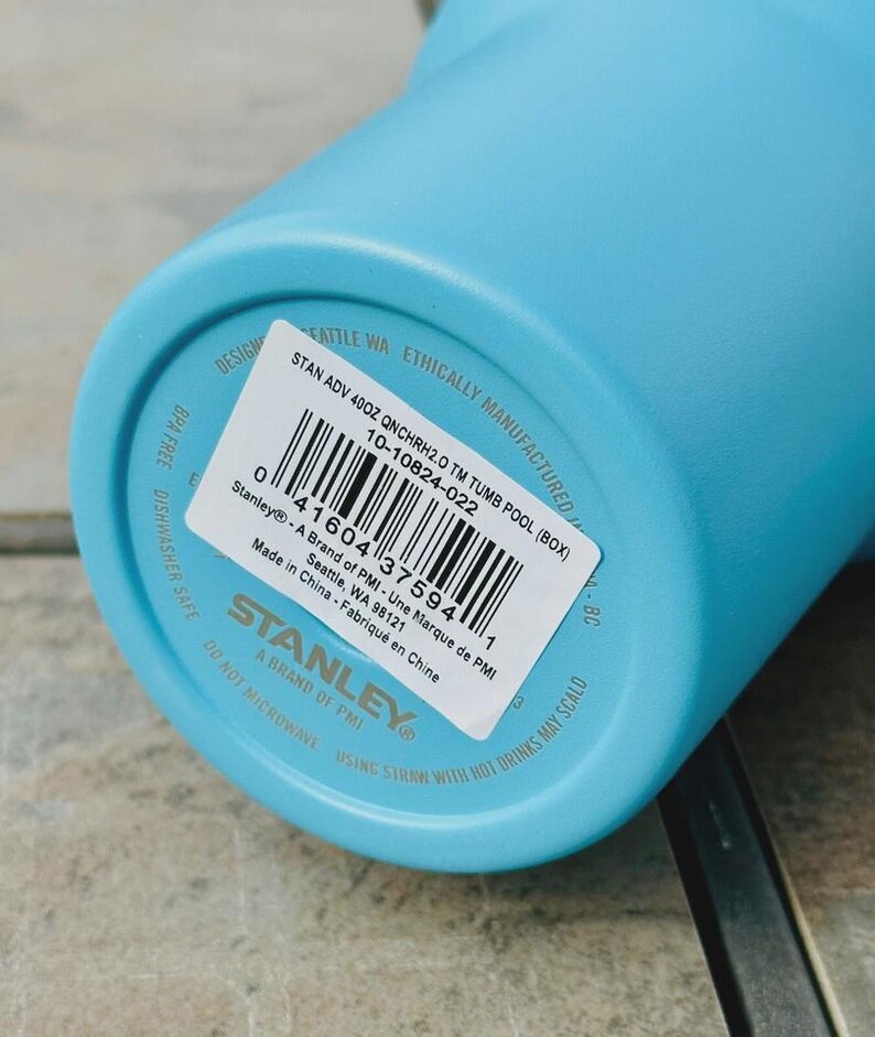 May include: A light blue Stanley 40oz tumbler. The base of the tumbler features a white sticker with product details, including the text "STAN ADV 40OZ QNCHRH2.0 TM TUMB POOL (BOX)" and a barcode. The tumbler is BPA free and dishwasher safe.
