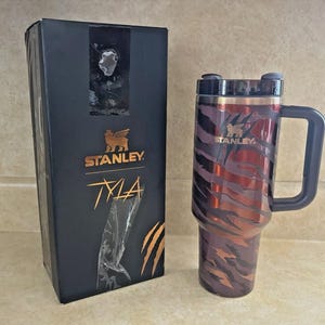 May include: A Stanley Tyla travel mug with a handle and a black lid, featuring a tiger stripe design in shades of red and brown. The mug is next to a black box with the Stanley logo and the word "Tyla" in gold.