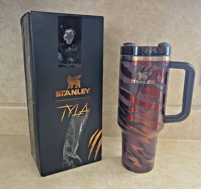 May include: A Stanley Tyla travel mug with a handle and a black lid, featuring a tiger stripe design in shades of red and brown. The mug is next to a black box with the Stanley logo and the word "Tyla" in gold.