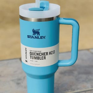 May include: A light blue Stanley Quencher H2.0 tumbler with a white lid and handle. The tumbler features the Stanley logo and text reading "Quencher H2.0 Tumbler" and "40 OZ | 1.18L". Includes a white straw.