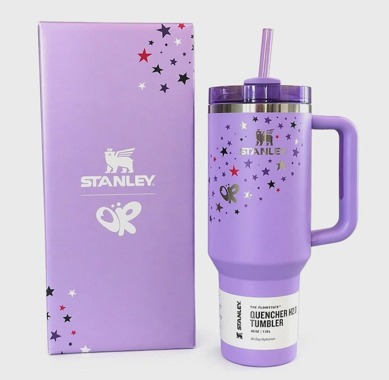 Op de afbeelding: Een lavendelkleurige Stanley Quencher H2.0 tumbler met een handvat, rietje en deksel, naast een bijpassende doos. De tumbler en doos zijn versierd met sterren en een vlinder. De tumbler bevat 1,18L.