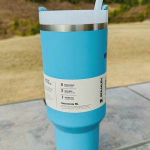 May include: A light blue Stanley tumbler with a white lid and a clear straw. The tumbler has a silver band and a label indicating it keeps drinks cold for 11 hours and iced for 2 days. The tumbler is 24 cm tall.