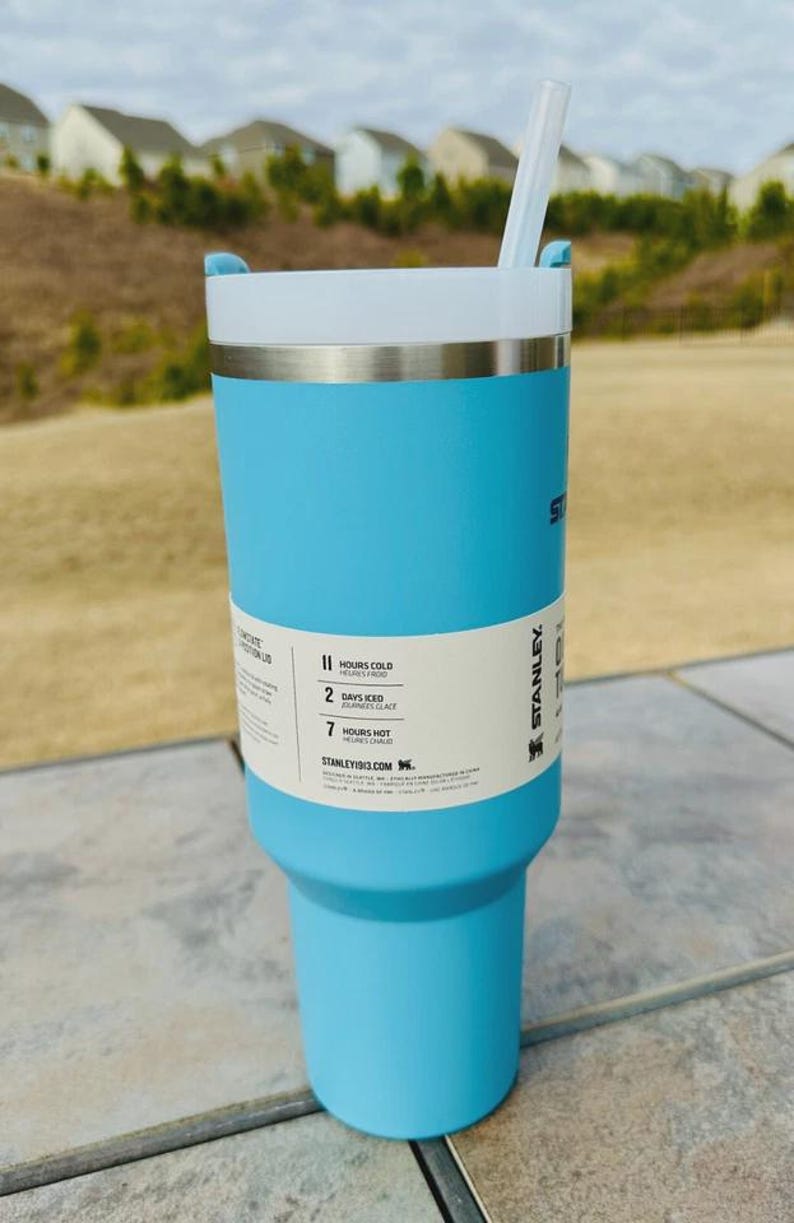 May include: A light blue Stanley tumbler with a white lid and a clear straw. The tumbler has a silver band and a label indicating it keeps drinks cold for 11 hours and iced for 2 days. The tumbler is 24 cm tall.