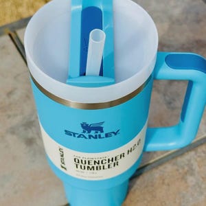 May include: A light blue Stanley Quencher H2.0 tumbler with a white interior and a matching handle. The tumbler features a straw and a lid with a sliding closure. The text "STANLEY" and "QUENCHER TUMBLER" are visible on the side.