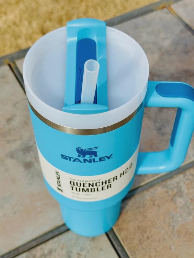 May include: A light blue Stanley Quencher H2.0 tumbler with a white interior and a matching handle. The tumbler features a straw and a lid with a sliding closure. The text "STANLEY" and "QUENCHER TUMBLER" are visible on the side.