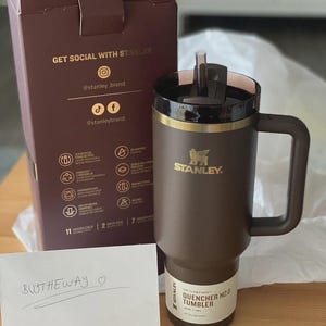 Peut inclure: Un gobelet Stanley Quencher H2.0 marron avec un couvercle et une poign&eacute;e noirs. Le gobelet a une bande dor&eacute;e et le logo Stanley. La bo&icirc;te derri&egrave;re dit "Get Social With Stanley". Une note manuscrite dit "BUTHTHEWAY".