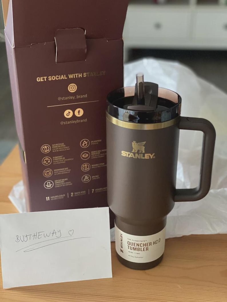 Peut inclure: Un gobelet Stanley Quencher H2.0 marron avec un couvercle et une poign&eacute;e noirs. Le gobelet a une bande dor&eacute;e et le logo Stanley. La bo&icirc;te derri&egrave;re dit "Get Social With Stanley". Une note manuscrite dit "BUTHTHEWAY".